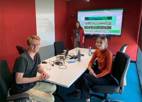 Three people in a meeting room facing the camera. On the far wall is a TV display with artworks.