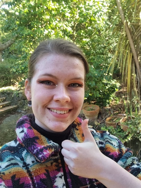 A young person with colourful clothing smiles and gives a thumbs up, with a garden in the background.